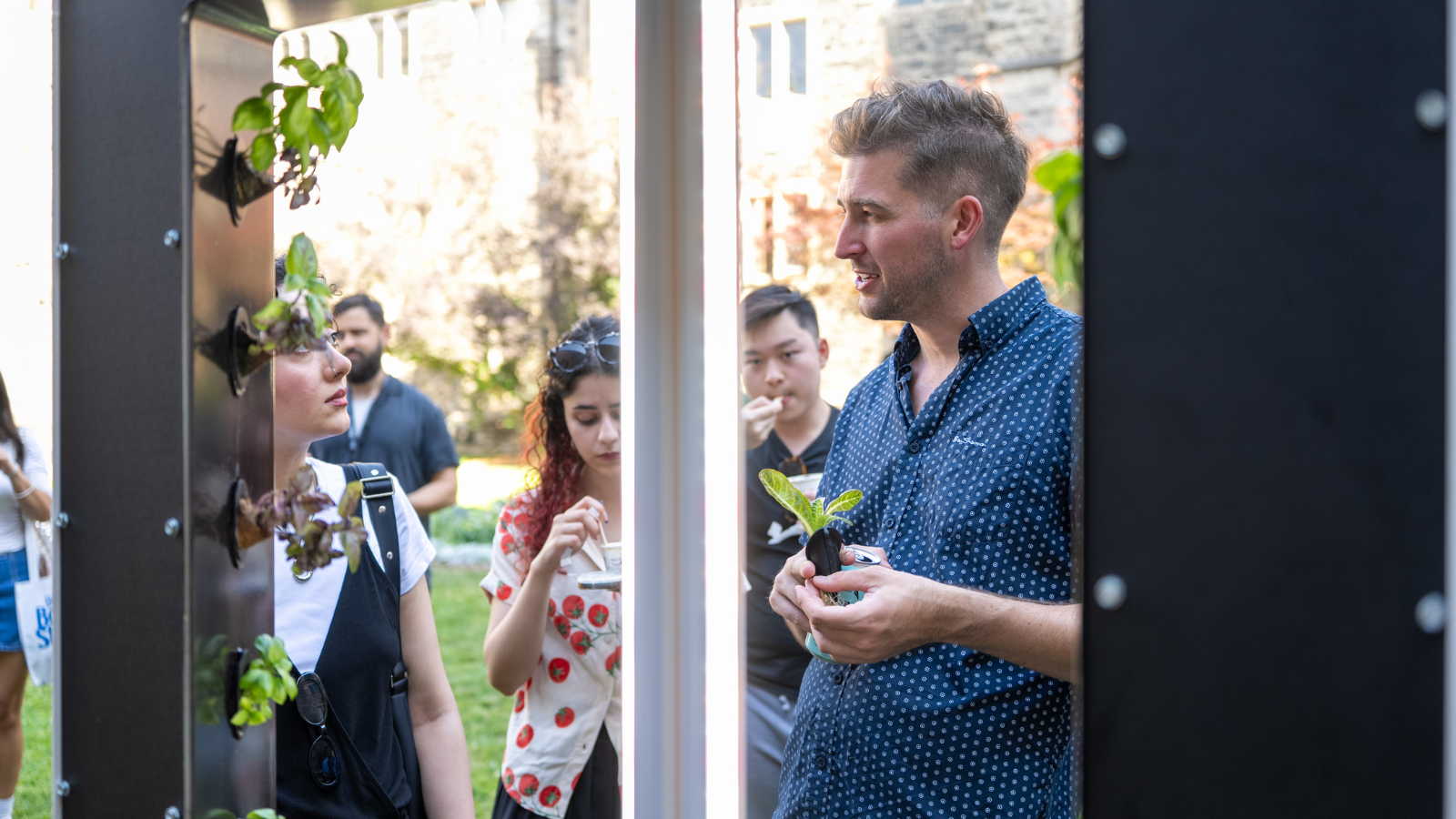 Conner Tidd holding a small hydroponic plant seedling, speaking to a group of interested people gathered outdoors around a modern vertical garden display unit.