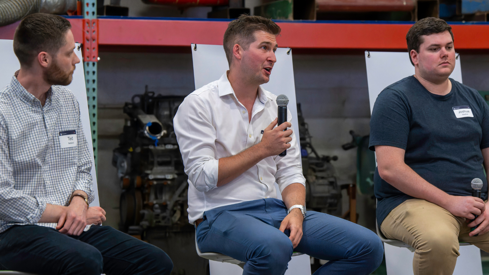 Three men seated on stage for a panel event. Conner Tidd, CEO and Co-Founder of Just Vertical, holds a microphone, and speaks actively while his co-panelists listen attentively.